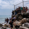 Crowded group of people on a rocky shoreline beside a chain-link fence, some entering the water and others watching from the rocks and fence edge.
