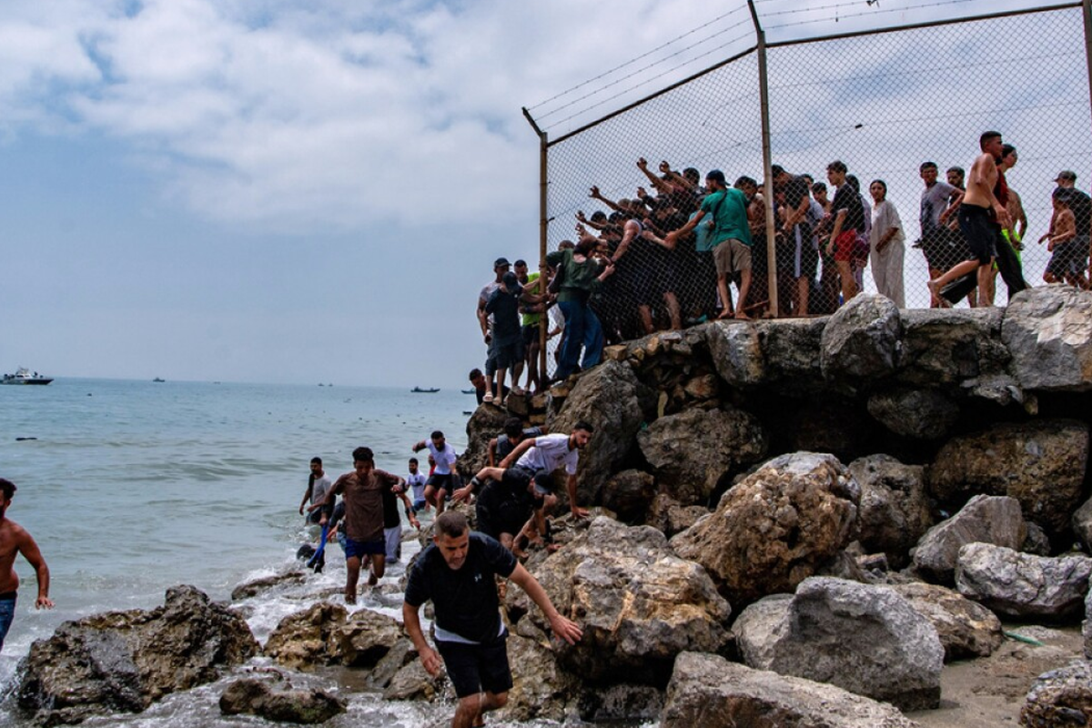 Crowded group of people on a rocky shoreline beside a chain-link fence, some entering the water and others watching from the rocks and fence edge.