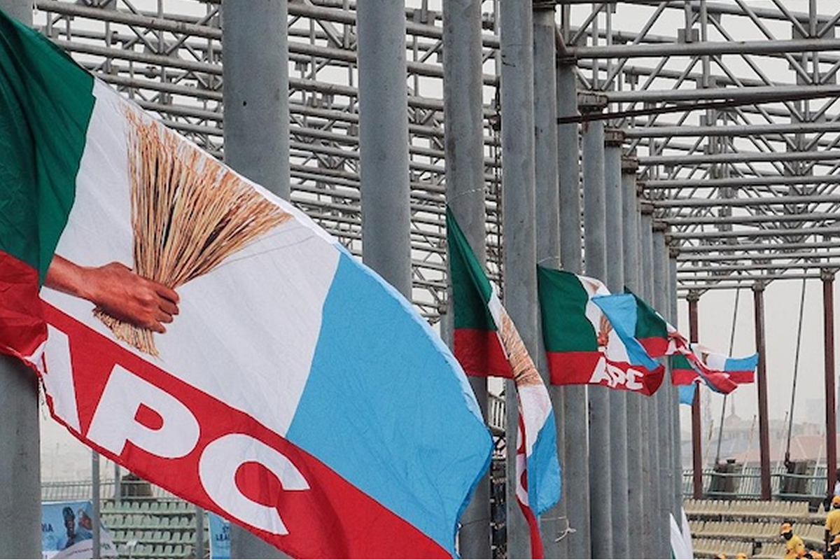 Flags in green, white, blue and red flutter along a metal framework at an outdoor event, with APC logos visible on some banners.