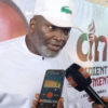 Man in a white outfit and cap speaks to reporters, microphone held near him and a smartphone recording nearby in an interview setting with a backdrop banner