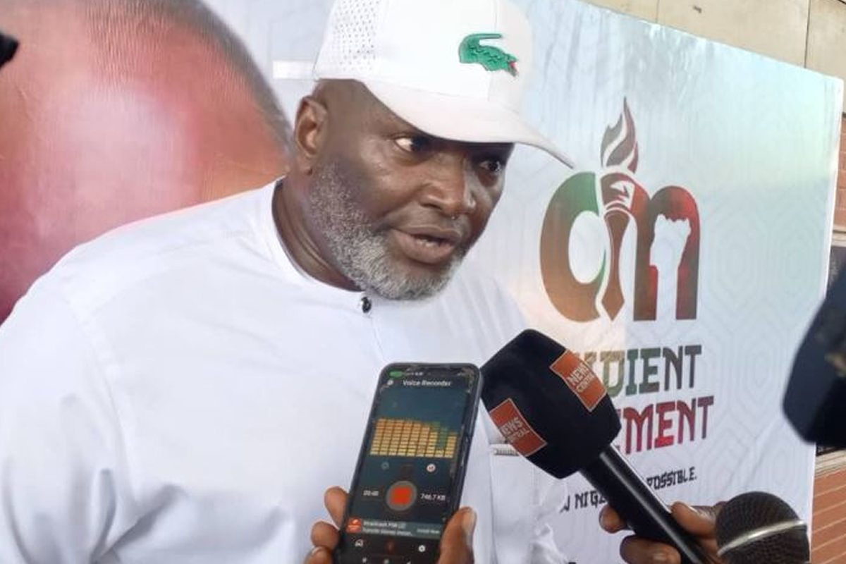 Man in a white outfit and cap speaks to reporters, microphone held near him and a smartphone recording nearby in an interview setting with a backdrop banner