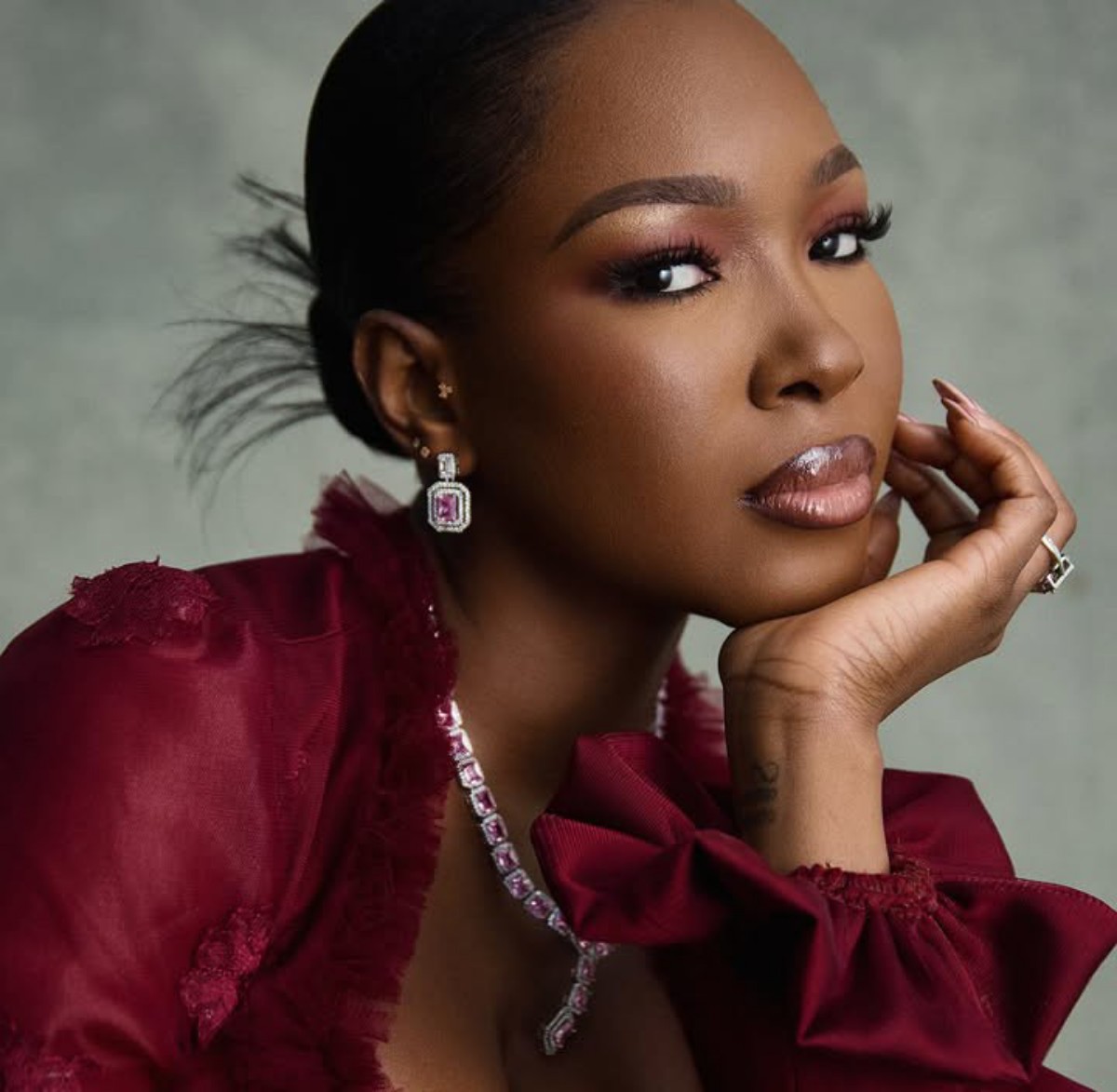 Close-up portrait of a woman with sleek hair, wearing diamond earrings and a matching necklace, in a burgundy lace dress, resting her chin on her hand.