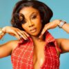 Glamour portrait of a woman with dark, voluminous hair and bold makeup, wearing a red checkered halter top and jewelry, posing with hands framing her face against a blue background.