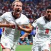 Two England footballers in white kits celebrate a goal on the pitch, crowd in the background, one wearing captain's armband on his left arm, both shouting with fists clenched.