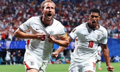Two England footballers in white kits celebrate a goal on the pitch, crowd in the background, one wearing captain's armband on his left arm, both shouting with fists clenched.