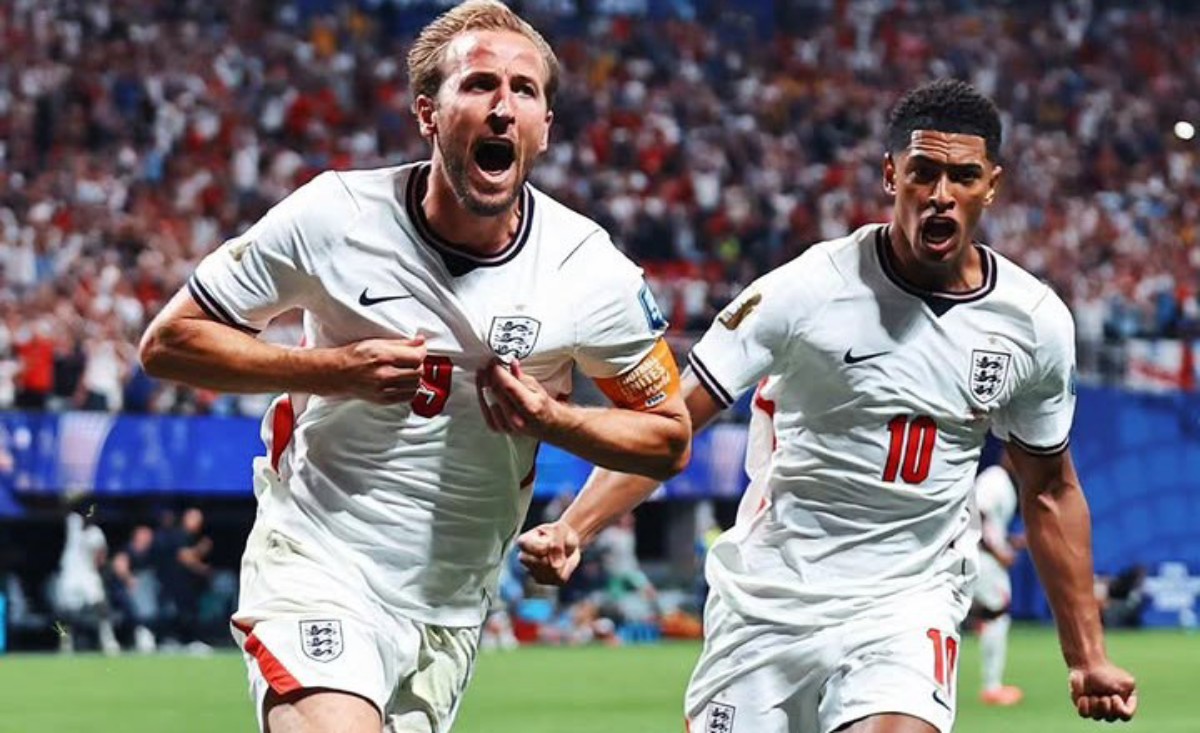 Two England footballers in white kits celebrate a goal on the pitch, crowd in the background, one wearing captain's armband on his left arm, both shouting with fists clenched.