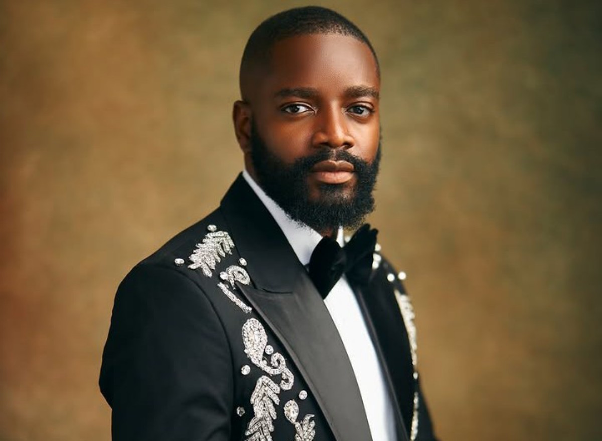 Portrait of a man in a black tuxedo with ornate silver embroidery, white shirt and bow tie, posed against a warm brown studio backdrop.