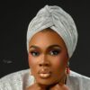 Portrait of a woman with a silver patterned headwrap and matching outfit, wearing gold earrings against a dark backdrop.
