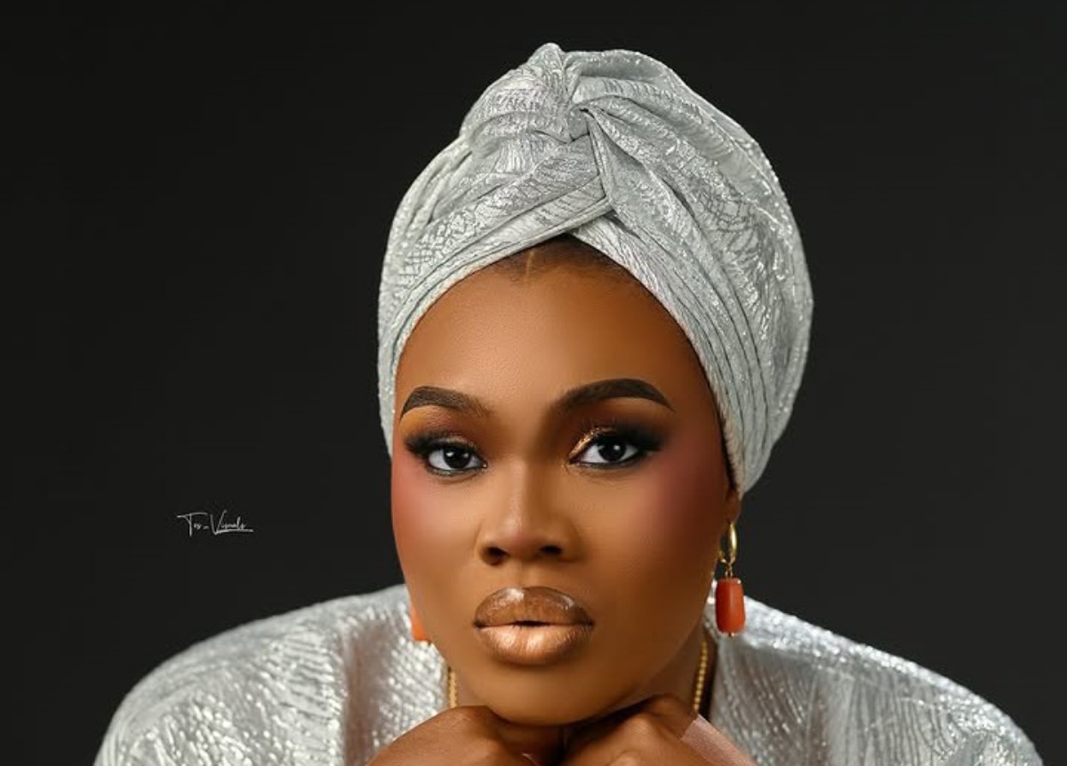 Portrait of a woman with a silver patterned headwrap and matching outfit, wearing gold earrings against a dark backdrop.