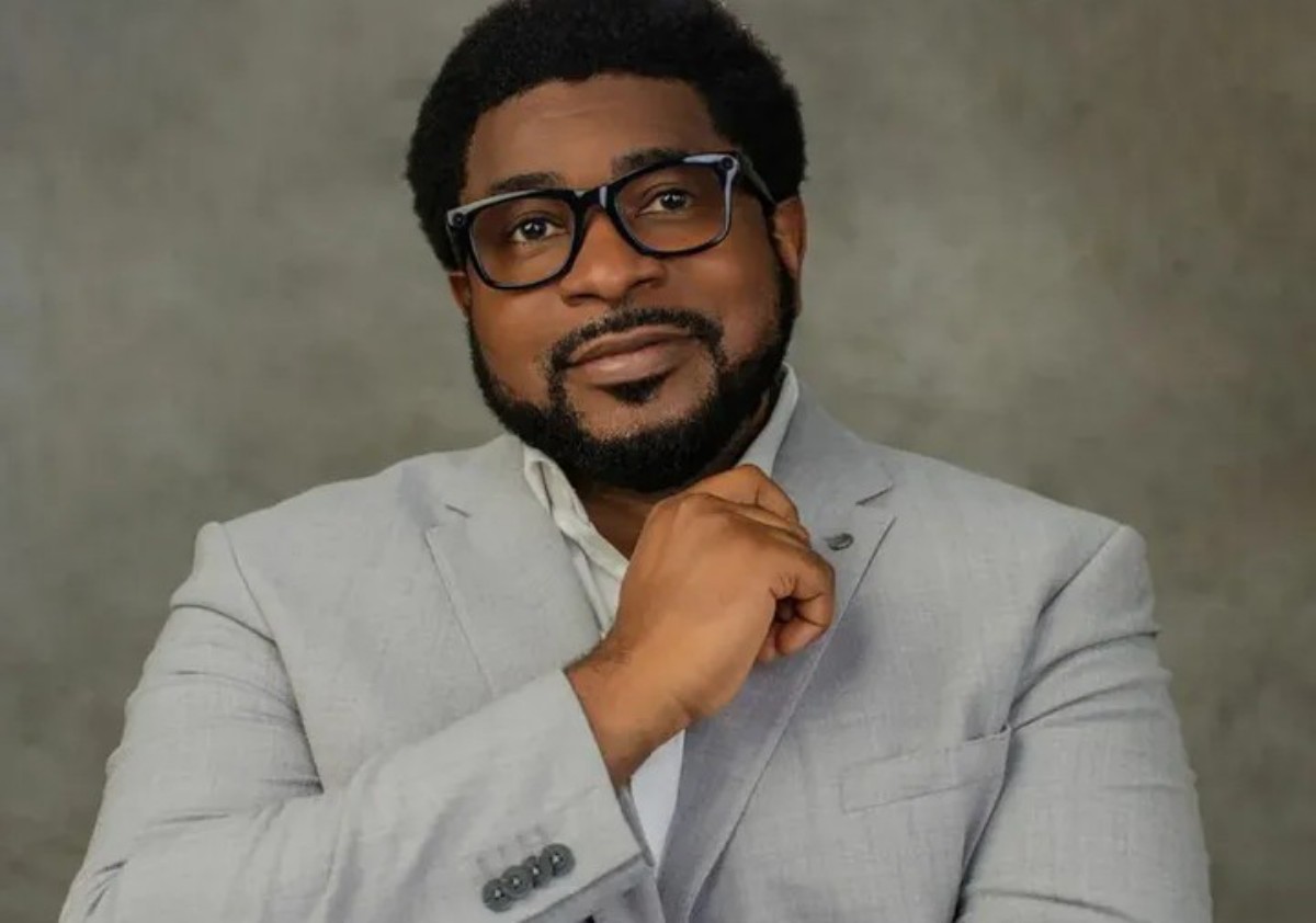 Portrait of a man wearing a light gray suit and white shirt, with black-ramed glasses and a short beard, looking at the camera with a thoughtful smile.