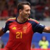 Soccer player in a red Spain jersey #21 celebrates on the field, pointing with his right arm toward the right side.