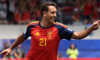 Soccer player in a red Spain jersey #21 celebrates on the field, pointing with his right arm toward the right side.