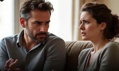 A man and woman sit on a sofa, facing each other with tense expressions during a heated discussion in a living room of a home.