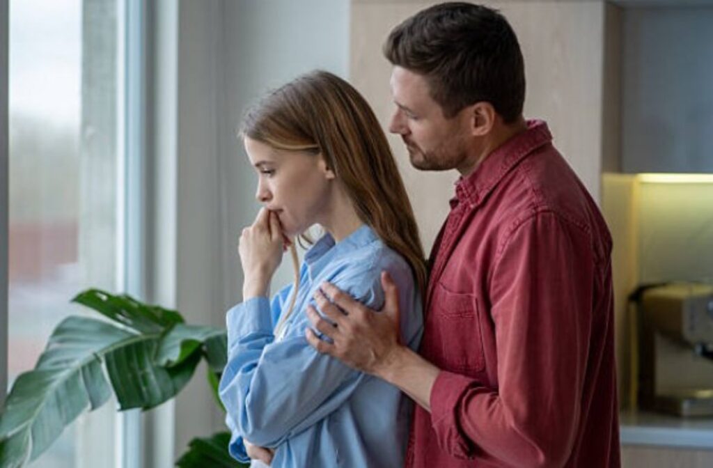 Woman with hand to her mouth, looking thoughtful, as a man places a reassuring hand on her shoulder in a bright home setting.
