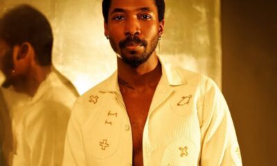 Portrait of a man with short hair and a trimmed beard, wearing a cream shirt with embroidered symbols, standing against a warm gold background.