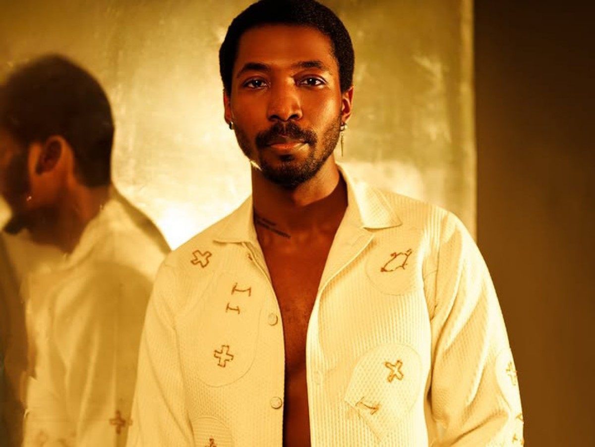 Portrait of a man with short hair and a trimmed beard, wearing a cream shirt with embroidered symbols, standing against a warm gold background.