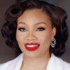 Close-up of a smiling woman with vintage curls, bold red lips, dramatic eyeliner, and sparkly earrings.