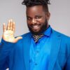 Smiling man in a bright blue suit waves hello with his right hand.