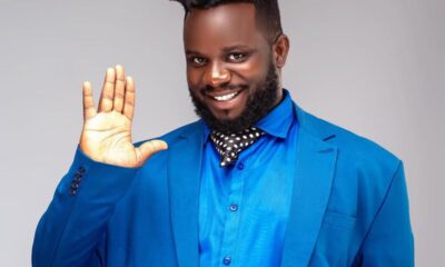 Smiling man in a bright blue suit waves hello with his right hand.