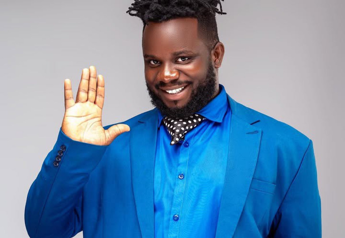 Smiling man in a bright blue suit waves hello with his right hand.