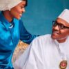 Woman in a blue traditional outfit smiles at an elderly man in white attire and cap, against a teal backdrop.