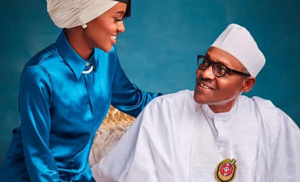 Woman in a blue traditional outfit smiles at an elderly man in white attire and cap, against a teal backdrop.