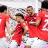 Four soccer players in red jerseys celebrate a goal near the goal net, high-fiving and smiling.