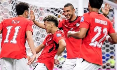 Four soccer players in red jerseys celebrate a goal near the goal net, high-fiving and smiling.