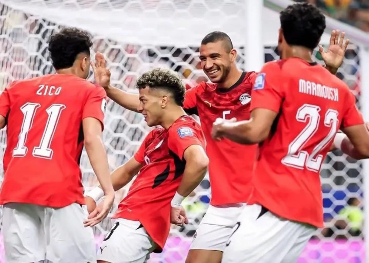 Four soccer players in red jerseys celebrate a goal near the goal net, high-fiving and smiling.