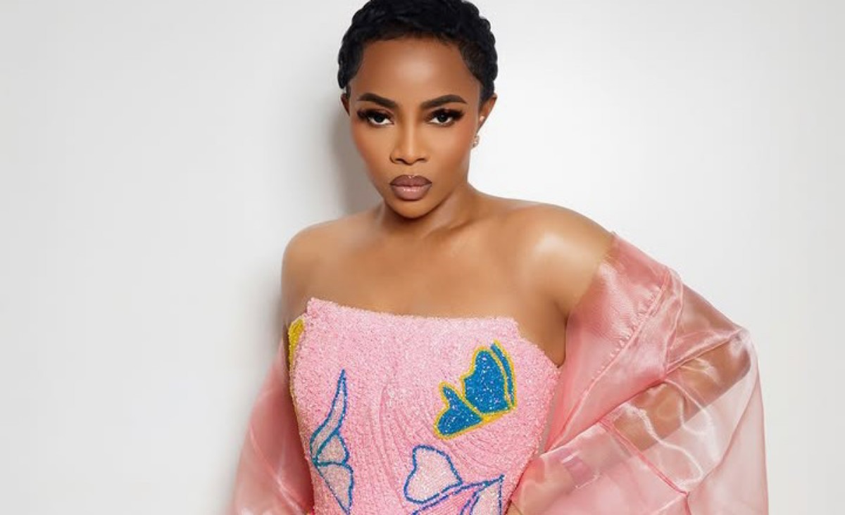 Portrait of a woman with short hair wearing a pink strapless dress with blue butterfly embellishments and a sheer pink shawl against a light background.