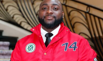 Portrait of a man with a beard wearing a red jacket with the number 14, a white shirt, and black tie, indoors by a curved railing.