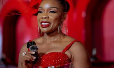 Smiling woman in a red sequined dress holds a microphone on a red-lit stage backdrop.