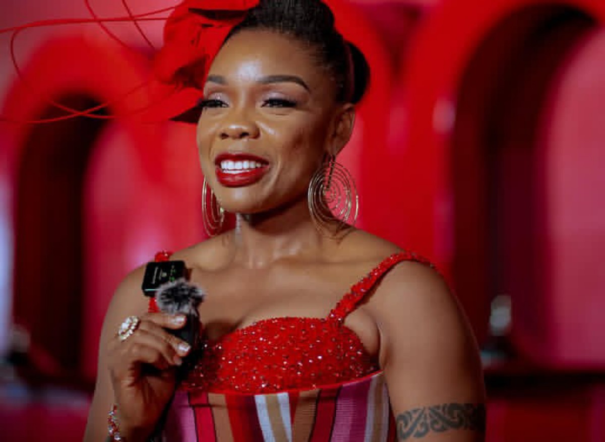 Smiling woman in a red sequined dress holds a microphone on a red-lit stage backdrop.
