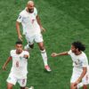 Three Moroccan football players in white jerseys celebrate on a green pitch, one jumps mid-air while others reach out in triumph.