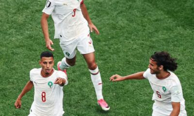 Three Moroccan football players in white jerseys celebrate on a green pitch, one jumps mid-air while others reach out in triumph.