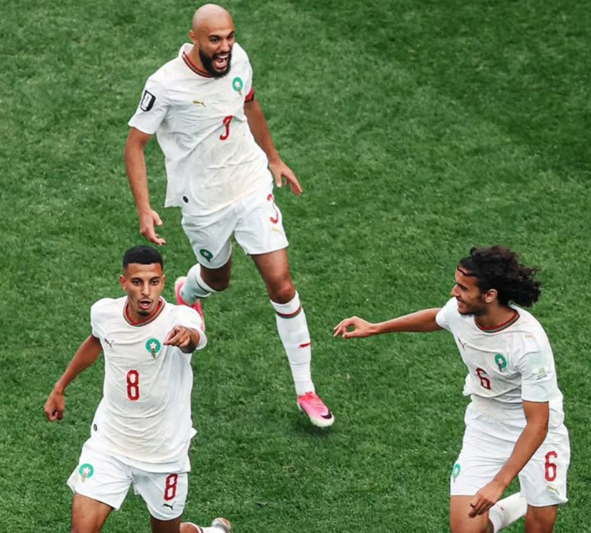 Three Moroccan football players in white jerseys celebrate on a green pitch, one jumps mid-air while others reach out in triumph.