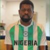 Man with beard wearing a Nigeria national football team jersey indoors, looking at the camera.