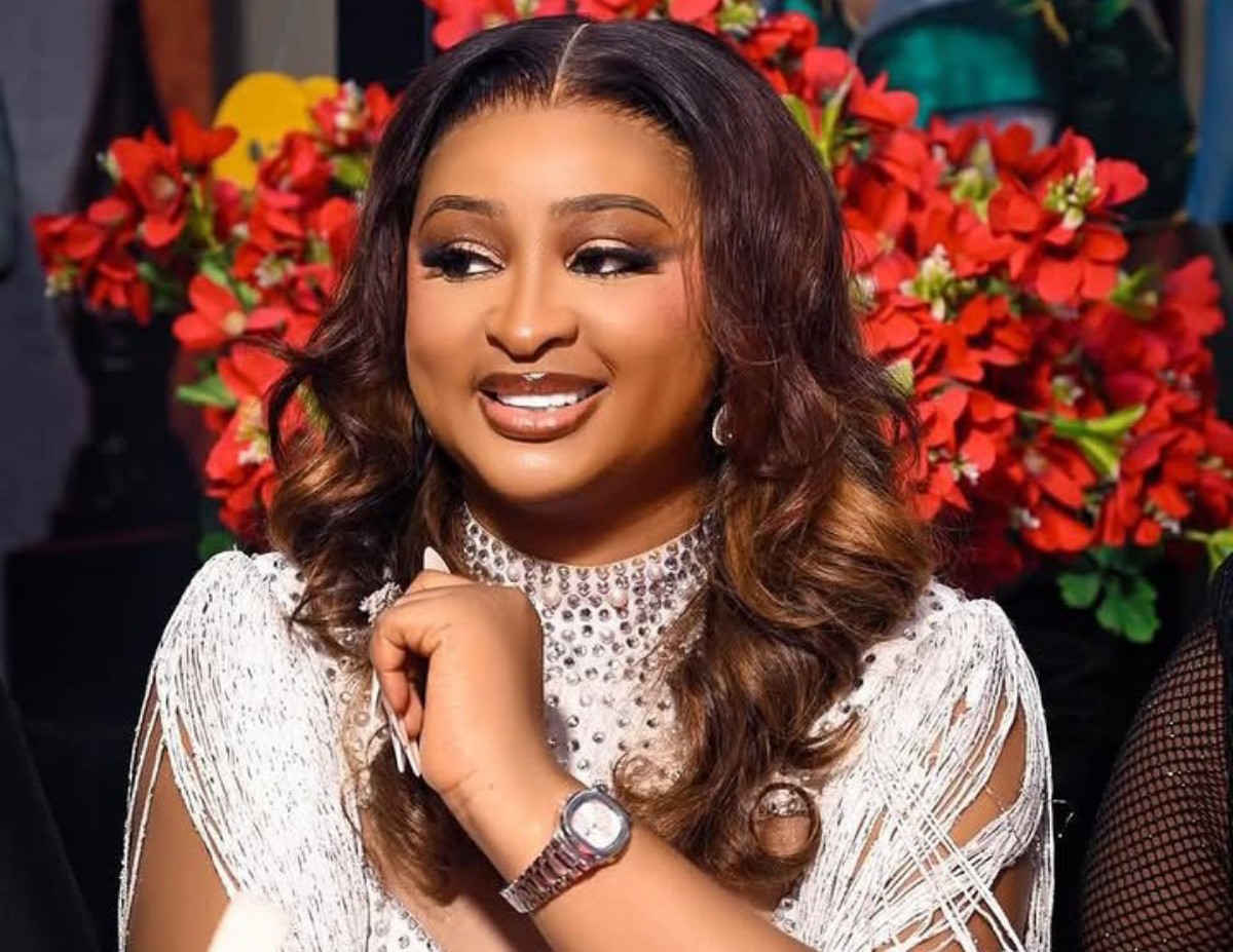 Smiling woman with wavy hair wearing a jeweled high-neck top and a wristwatch, posed in front of red flowers.