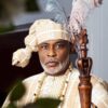 Portrait of a man in ceremonial royal attire with a feathered headdress and beaded necklace, holding a carved wooden staff.