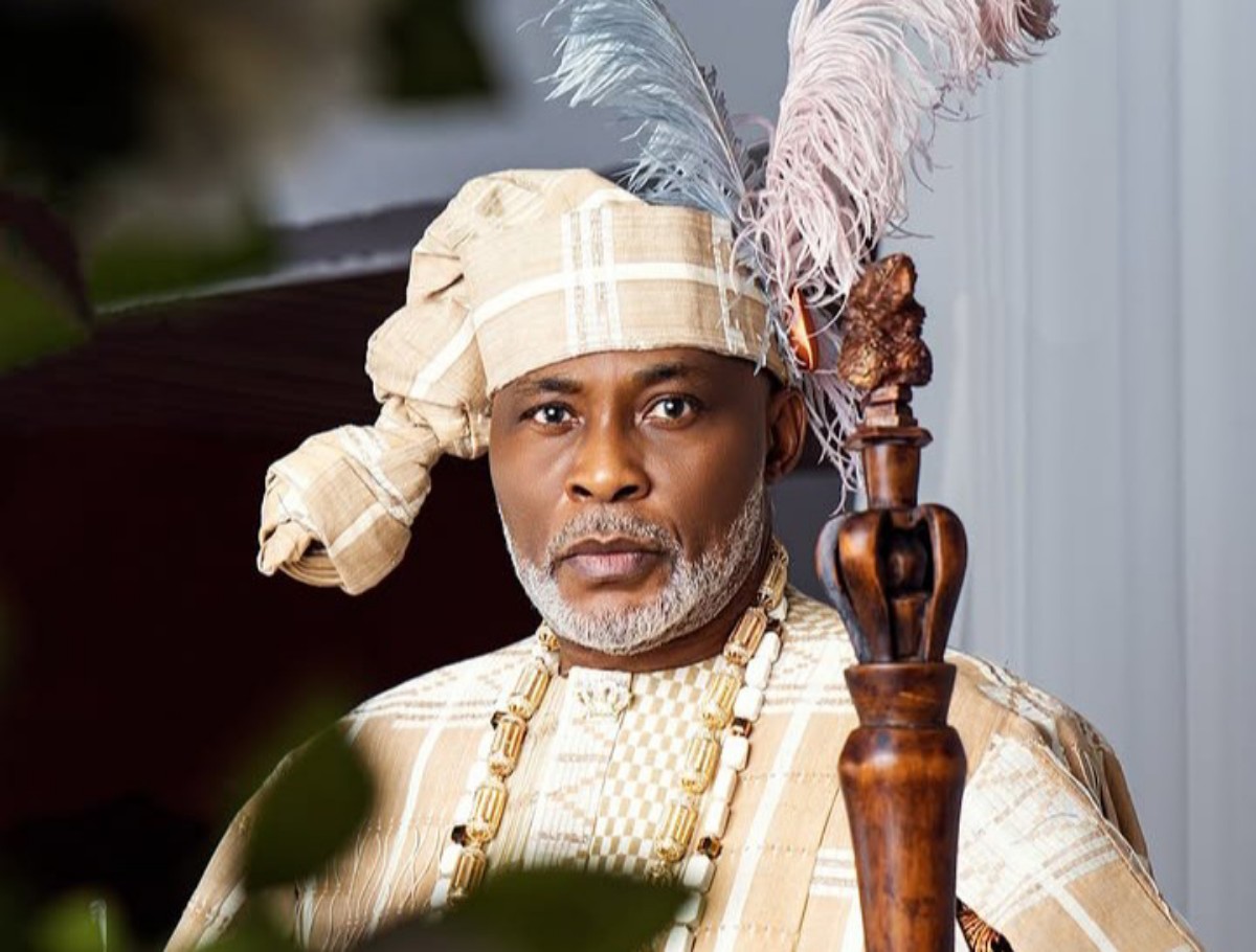 Portrait of a man in ceremonial royal attire with a feathered headdress and beaded necklace, holding a carved wooden staff.