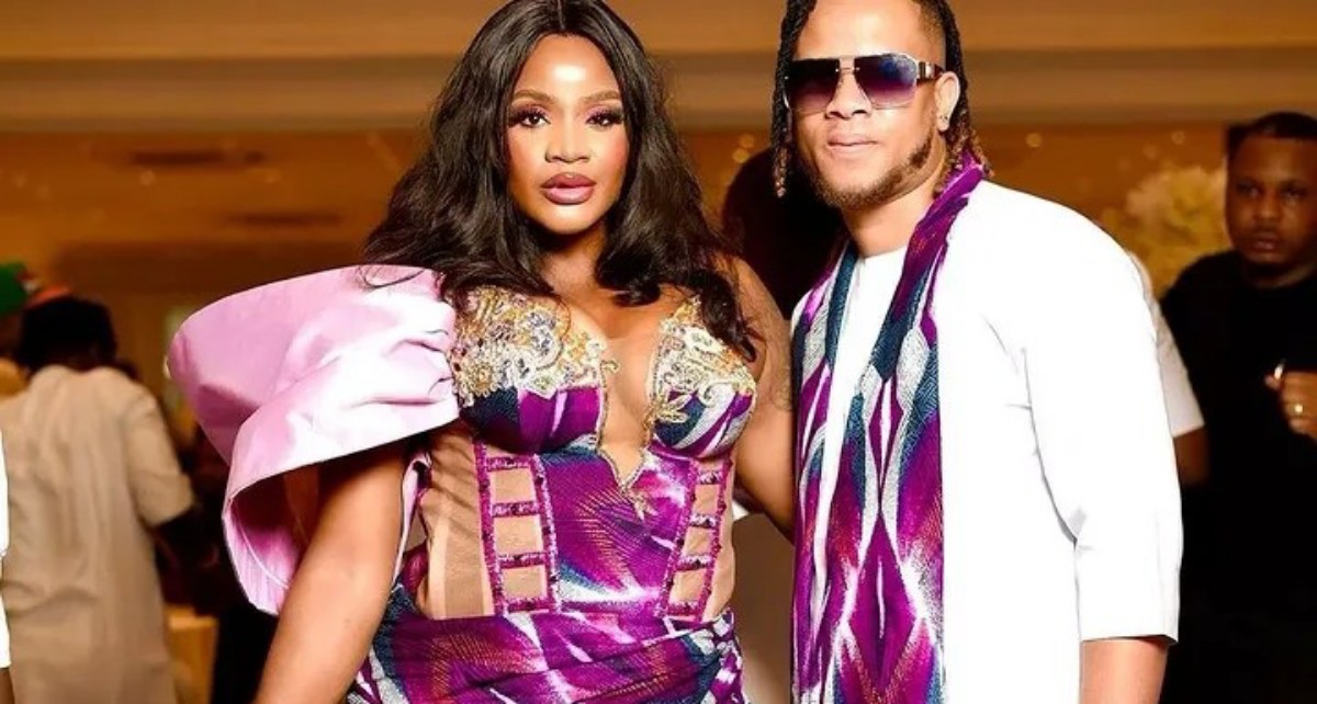 Two stylishly dressed people posing for a photo at a party, the woman wearing a purple embellished gown with a pink cape and the man in a white outfit with a purple scarf and sunglasses.