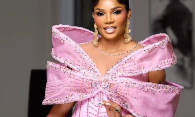 Woman in a pink off-shoulder gown with a large jeweled bow and gold earrings, posing indoors for a photo.