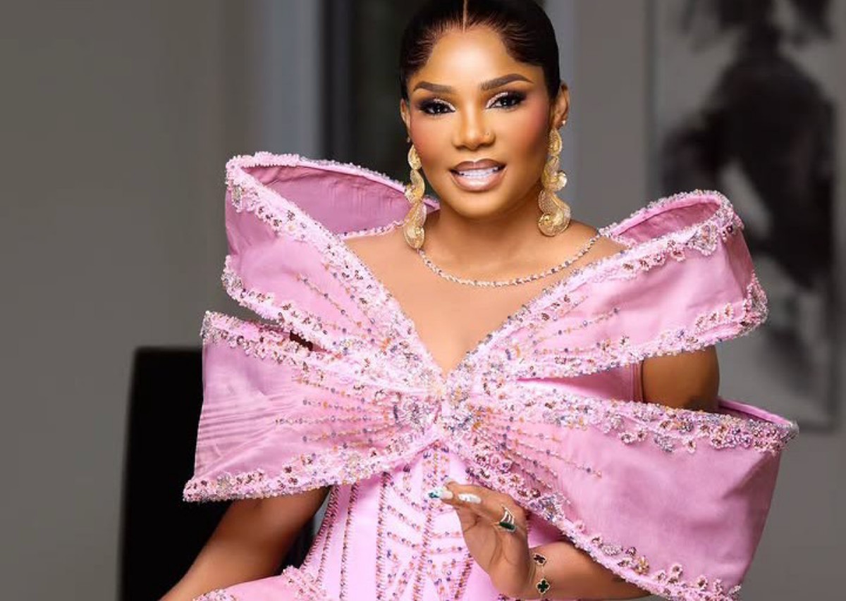 Woman in a pink off-shoulder gown with a large jeweled bow and gold earrings, posing indoors for a photo.