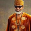 Man in orange traditional attire with a patterned hat, sunglasses, and a beaded necklace, smiling at the camera.