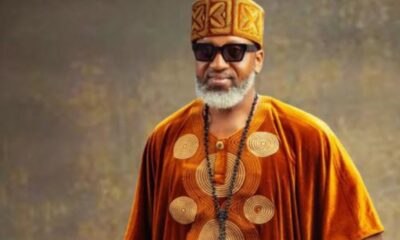 Man in orange traditional attire with a patterned hat, sunglasses, and a beaded necklace, smiling at the camera.
