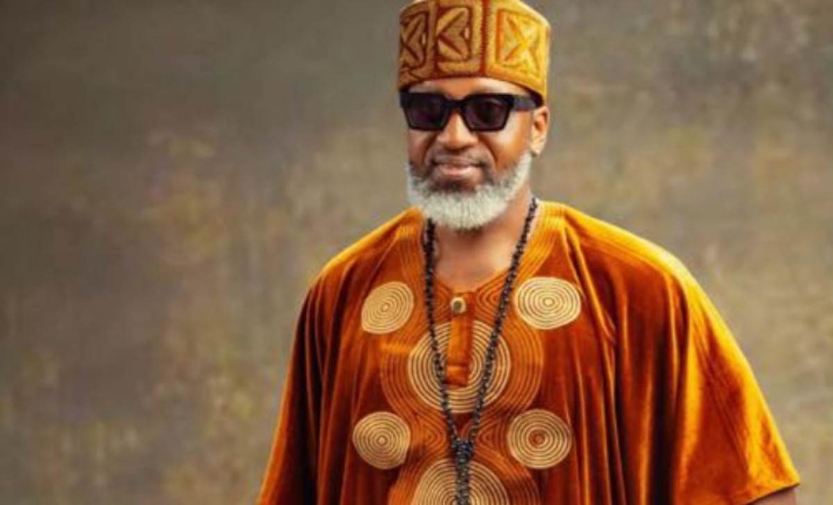 Man in orange traditional attire with a patterned hat, sunglasses, and a beaded necklace, smiling at the camera.
