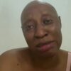 Close-up of a person with a shaved head looking at the camera, wearing a pink sleeveless top, with a calm expression.
