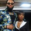 Man with a bearded face wearing a glittery blue-green sequin blazer, sunglasses, and gold jewelry stands beside a woman in a black blazer with white lapels near a car in a parking area.