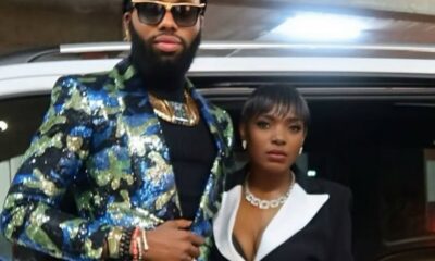 Man with a bearded face wearing a glittery blue-green sequin blazer, sunglasses, and gold jewelry stands beside a woman in a black blazer with white lapels near a car in a parking area.