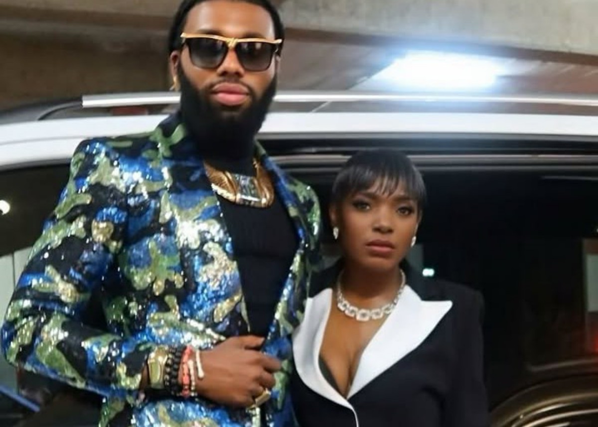 Man with a bearded face wearing a glittery blue-green sequin blazer, sunglasses, and gold jewelry stands beside a woman in a black blazer with white lapels near a car in a parking area.